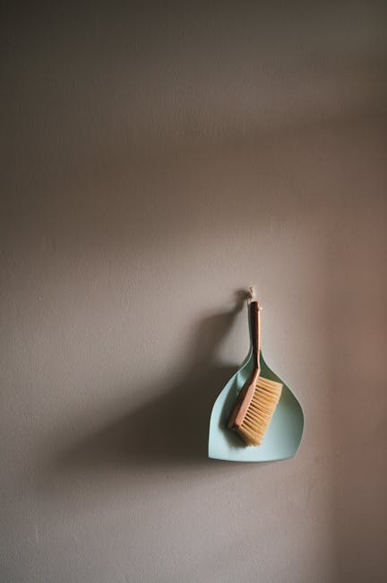 A light blue wall hook holding a wooden-handled brush with beige bristles and a matching dustpan with a wooden handle, both positioned to the right side of the image. The wall has a smooth, matte fini