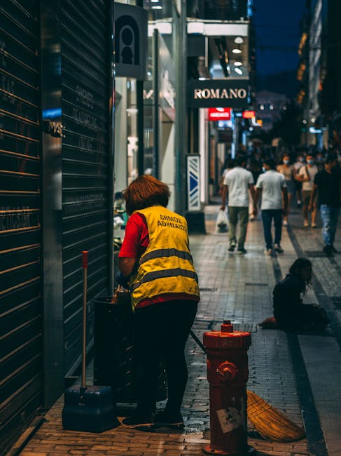 A street cleaner dressed in bright orange high-visibility clothing, including a jacket with reflective stripes and a hood, is sweeping the pavement with a red-bristled broom. The cleaner is standing n